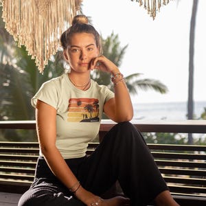 May include: A woman wearing a light green t-shirt with a sunset graphic and the words "Read, Tan, Repeat." She is sitting on a wooden bench, wearing black trousers, and has her hair in a bun. The background includes macrame decor and a beach view.