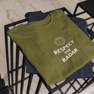 May include: Olive green t-shirt with the text "Respect the Radar" in white. The shirt is folded and placed on top of a navy blue t-shirt. The shirts are on a black metal shelf.