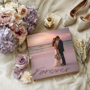Puede incluir: Un álbum de fotos de boda con una escena de playa y la palabra "Forever" escrita en la arena. El álbum está rodeado de un ramo de flores, un par de tacones adornados y un pequeño objeto decorativo.