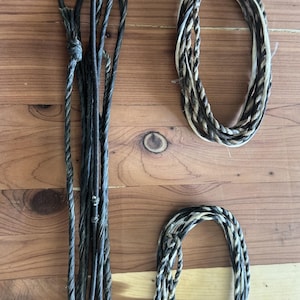 May include: An assortment of braided and twisted ropes in shades of black, gray, and tan. The ropes are arranged on a wooden surface, with some coiled into loops and others hanging straight down. The textures vary from smooth to tightly wound.