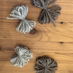 May include: Four handmade yarn pom-poms in shades of gray and brown are arranged on a wooden surface. The pom-poms are made from thick yarn and have a fluffy, textured appearance. The wooden surface has a warm, natural wood grain.