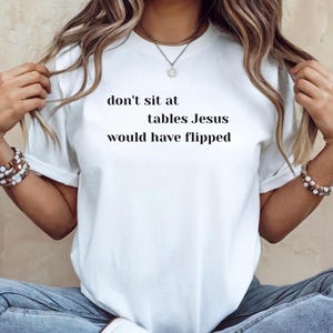 May include: White t-shirt with the text "don't sit at tables Jesus would have flipped" in black. The shirt is being worn with blue jeans and white sneakers. The person is wearing bracelets and a necklace.