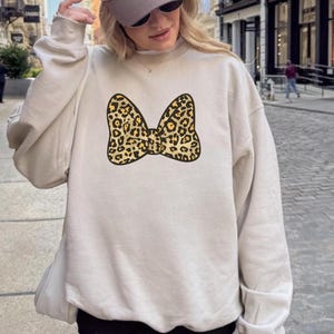 May include: A beige sweatshirt featuring a large bow design with a leopard print pattern. The bow is in shades of tan, brown, and black. The sweatshirt is a casual style, suitable for everyday wear.