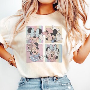 Puede incluir: Camiseta color crema con un dise&ntilde;o pop art colorido de Minnie Mouse en cuatro poses diferentes. Cada cuadrado muestra a Minnie con un lazo y una expresi&oacute;n diferentes, sobre fondos pastel.