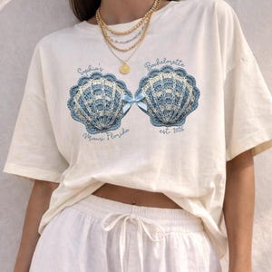 May include: Cream-colored t-shirt featuring a seashell design in blue and white crochet, with a blue bow. Text includes "Sophia's Bachelorette" and "Miami Florida est. 2026". The shirt is part of a bridal party theme.