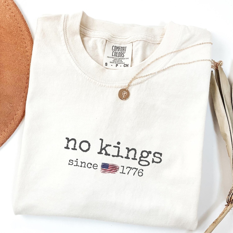 May include: Cream-colored t-shirt with the text "no kings since 1776" in black. The American flag is incorporated into the design. A gold necklace with a pendant rests on the shirt.