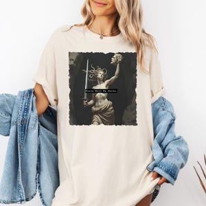 Puede incluir: Camiseta color crema con un gr&aacute;fico de una estatua cl&aacute;sica de una mujer sosteniendo una espada y una cabeza cortada. El texto "Girls Will Be Girls" est&aacute; impreso debajo de la imagen. La camiseta tiene mangas largas y un corte holgado.