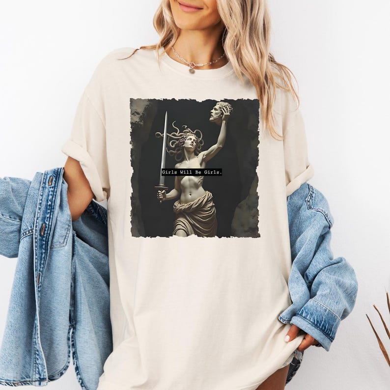 Puede incluir: Camiseta color crema con un gr&aacute;fico de una estatua cl&aacute;sica de una mujer sosteniendo una espada y una cabeza cortada. El texto "Girls Will Be Girls" est&aacute; impreso debajo de la imagen. La camiseta tiene mangas largas y un corte holgado.