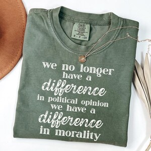 May include: An olive green t-shirt with the text "we no longer have a difference in political opinion we have a difference in morality" in white. A gold necklace with a small pendant is on the shirt.