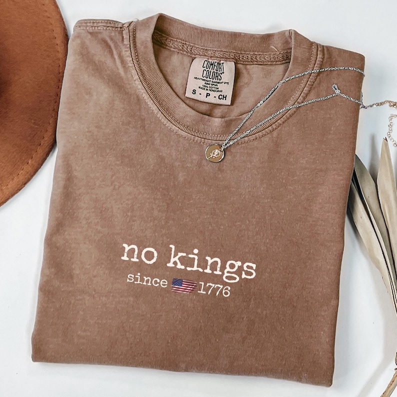 May include: A brown Comfort Colors t-shirt with the words "no kings since 1776" printed in white. A silver necklace with a small pendant is draped across the shirt. A brown hat is visible in the upper left corner.