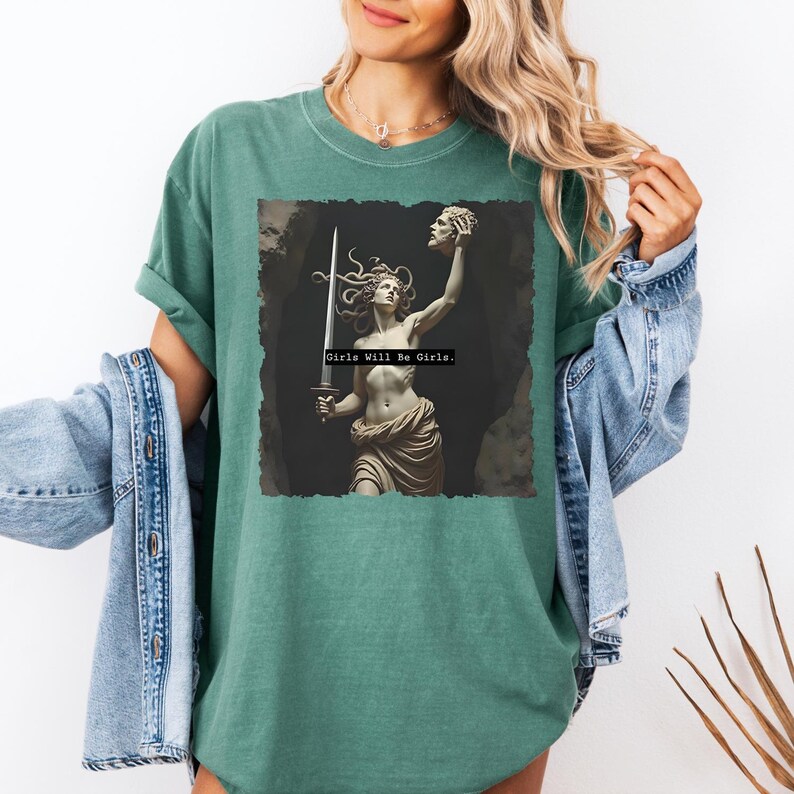 Puede incluir: Camiseta de color verde azulado con un gr&aacute;fico de una estatua cl&aacute;sica de una mujer sosteniendo una espada y una cabeza cortada. El texto "Girls Will Be Girls" est&aacute; impreso debajo. La camiseta tiene mangas cortas y un corte holgado.