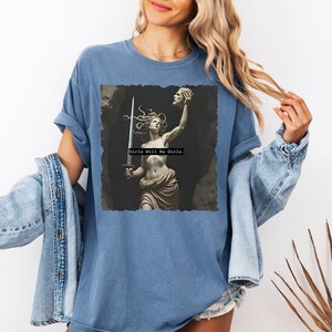 Puede incluir: Una camiseta azul con un gr&aacute;fico de una estatua cl&aacute;sica de una mujer sosteniendo una espada y una cabeza cortada. El texto "Girls Will Be Girls" est&aacute; impreso debajo de la imagen. La camiseta tiene mangas cortas y un corte relajado.