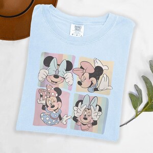 Puede incluir: Camiseta azul claro con un gr&aacute;fico colorido de Minnie Mouse en cuatro poses diferentes. Cada imagen de Minnie est&aacute; sobre un fondo de colores pastel. La camiseta est&aacute; doblada cuidadosamente, mostrando el dise&ntilde;o.