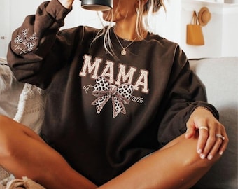 May include: A brown sweatshirt with the word "MAMA" in white block letters, a leopard print bow, and the year "2026". The sweatshirt also features a leopard print bow on the left sleeve.
