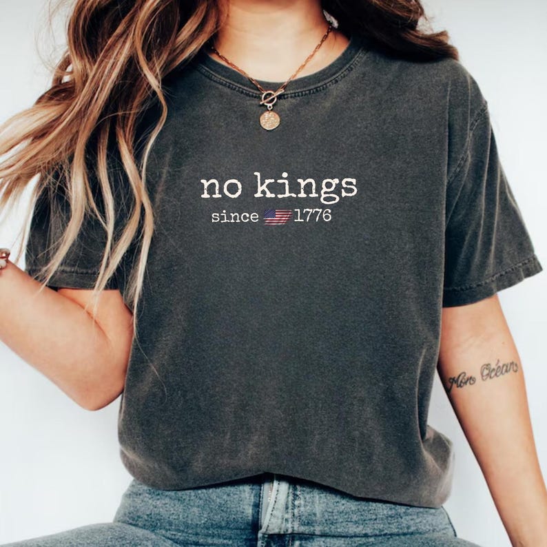 May include: Dark gray t-shirt with the text "no kings" above "since 1776" and an American flag graphic. The shirt is a short-sleeved, crew neck style.