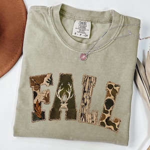 May include: A sage green short-sleeve t-shirt with a crew neckline and a Comfort Colors brand label. The front features the word FALL in large block letters, each with a unique pattern including camouflage, a deer antler, and tree bark.