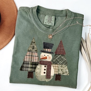 May include: Sage green Comfort Colors t-shirt with a snowman and Christmas tree applique. The snowman has a black top hat, orange carrot nose, and a brown scarf. The trees are made of plaid fabric. A silver necklace is draped across the shirt.