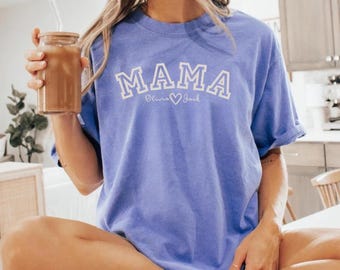 May include: A periwinkle blue t-shirt with the word "MAMA" in white block letters. Below the word "MAMA" is the text "Olivia ♡ Jack". The shirt is a relaxed fit and has short sleeves.