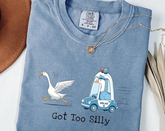 May include: A blue Comfort Colors t-shirt with a whimsical design. It features a cartoon goose running from a police car driven by a goose. The text "Got Too Silly" is printed below the graphic. A gold necklace is draped across the shirt.