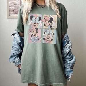 Puede incluir: Camiseta verde oliva con un gr&aacute;fico de cuatro paneles de Minnie Mouse en varias poses. Cada panel tiene un fondo pastel con rayas verticales. Minnie lleva diferentes lazos y atuendos en cada panel. La camiseta tiene un corte holgado.