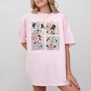 Puede incluir: Una camiseta rosa claro con un gr&aacute;fico de cuatro paneles de Minnie Mouse en varias poses. Cada panel tiene un fondo pastel. La camiseta tiene un corte holgado y de gran tama&ntilde;o.