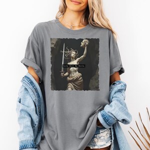 Puede incluir: Camiseta gris con un gr&aacute;fico de una estatua cl&aacute;sica de una mujer sosteniendo una espada y una cabeza cortada. El texto "Girls Will Be Girls" est&aacute; impreso debajo de la imagen. La camiseta tiene mangas enrolladas.