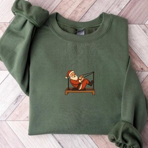 May include: A green sweatshirt featuring an embroidered Santa Claus exercising on a Pilates reformer. Santa is wearing his traditional red suit and hat. The sweatshirt is a solid, muted green color.