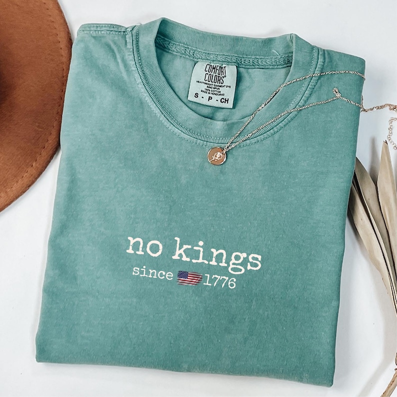May include: A teal Comfort Colors t-shirt with the words "no kings" and "since 1776" printed in white, along with a small American flag graphic. A gold necklace with a pendant rests on the shirt.