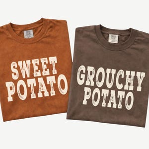 May include: Two crew neck t-shirts with Comfort Colors labels. The left shirt is rust orange with the white text SWEET POTATO. The right shirt is dark brown with the white text GROUCHY POTATO. Both shirts feature a distressed, weathered font style.
