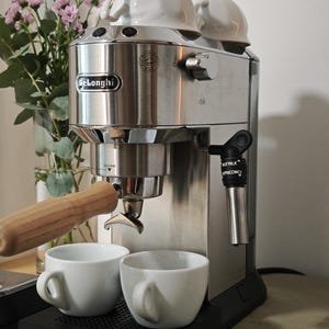 Può includere: Una macchina per espresso De'Longhi in acciaio inossidabile con portafiltro dal manico in legno, un erogatore di vapore nero con le scritte HOT MILK e CAPPUCCINO e un vassoio raccogligocce nero. Due tazze bianche poggiano sul vassoio, due capovolte.