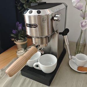 Op de afbeelding: Een roestvrijstalen De'Longhi espressomachine met een filterdrager met lichthouten handvat, een zwarte lekbak en een zilverkleurige stoompijp. Het apparaat heeft drie knoppen aan de bovenzijde, een logo aan de voorzijde en twee witte kopjes.