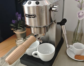 Peut inclure: Une machine à expresso De'Longhi en acier inoxydable avec un porte-filtre à poignée en bois clair, un plateau d'égouttage noir et une buse vapeur argentée. L'appareil comporte trois boutons supérieurs, un logo frontal et deux tasses blanches.