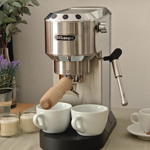 Könnte beinhalten: Eine De'Longhi Espressomaschine aus Edelstahl mit einem Siebträgergriff aus hellem Holz, einem schwarzen Sockel und einer silbernen Dampfdüse. Zwei weiße Keramiktassen stehen auf der Abtropfschale, eine dritte Tasse steht rechts auf einer Untertasse.