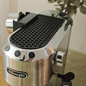 Könnte beinhalten: Eine silberne DeLonghi Espressomaschine mit einer schwarzen, wabenförmigen Ablagefläche, drei runden Bedientasten und einer seitlichen Dampfdüse mit der Aufschrift HOT MILK CAPPUCCINO. Das Gerät hat eine metallische Oberfläche und einen Siebträger.