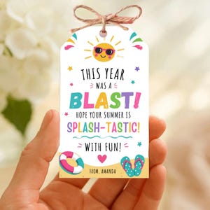 Puede incluir: Una etiqueta de regalo blanca con temática de verano, con un sol sonriente con gafas de sol y el texto "THIS YEAR WAS A BLAST!" en letras coloridas. La etiqueta incluye un corazón, un salvavidas y chanclas.