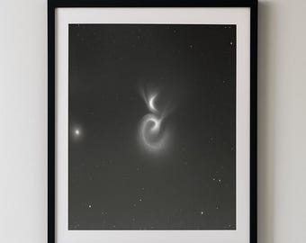 May include: Monochrome framed print depicting a galaxy with swirling patterns and a starry backdrop. The artwork is mounted on a white wall, above a grey sofa with a white cushion.