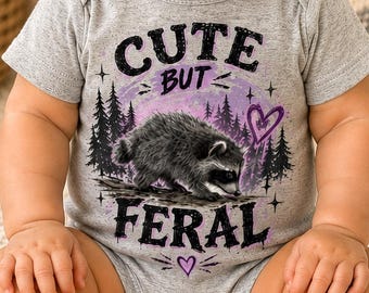 May include: A heather gray short-sleeve baby bodysuit featuring a graphic print of a raccoon in a forest setting. The text reads CUTE BUT FERAL in black lettering, accented by a purple heart and small star symbols, centered on the chest.