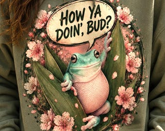 May include: An olive green hooded sweatshirt featuring a graphic print of a frog surrounded by pink cherry blossoms. The text "HOW YA DOIN', BUD?" appears in a speech bubble above the frog. The hoodie includes a front kangaroo pocket and drawstrings.