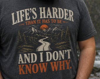 May include: A charcoal gray short-sleeve t-shirt featuring a graphic print of a winding road leading toward mountains and a sun. The text reads: LIFE'S HARDER THAN IT HAS TO BE AND I DON'T KNOW WHY. The shirt is paired with a light beige sun hat.