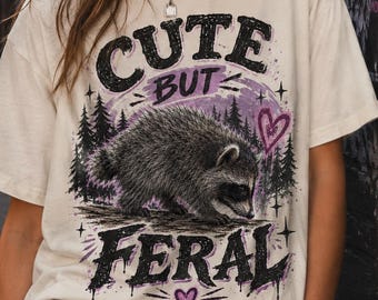 May include: An off-white short-sleeve t-shirt featuring a graphic print of a raccoon in a forest setting. The text reads CUTE BUT FERAL in bold black lettering, accented by a purple circular background, small stars, and two purple heart icons.