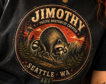 May include: A black short-sleeve t-shirt featuring a circular graphic with a raccoon illustration. The text reads JIMOTHY, PACIFIC NORTHWEST CRYPTID, and SEATTLE WA. The design includes a mountain, forest, and the Space Needle in a red and orange sunset.