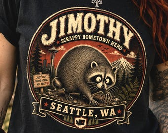 May include: A black t-shirt featuring a circular graphic of a raccoon with the text "JIMOTHY SCRAPPY HOMETOWN HERO", "JUST OUT HERE DOING MY BEST", and "SEATTLE, WA". The design includes a mountain, a city skyline, and a small state of Washington icon.