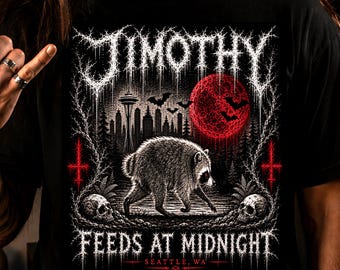 May include: A black t-shirt featuring a graphic print of a raccoon walking in a forest at night. The design includes a red moon, bats, a city skyline with the Space Needle, two skulls, and the text JIMOTHY FEEDS AT MIDNIGHT SEATTLE, WA.