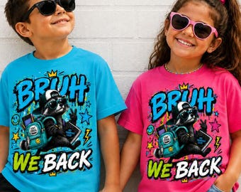 May include: A bright blue t-shirt and a bright pink t-shirt, each featuring a graphic of a raccoon wearing sunglasses and a cap, carrying a backpack, with the text "BRUH WE BACK" in bold blue and green letters, surrounded by stars and lightning bolts.
