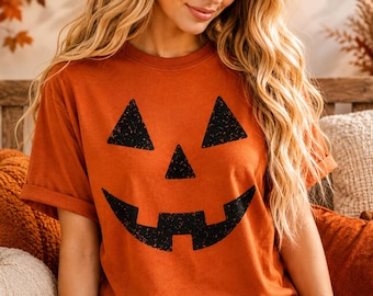 May include: A burnt orange short-sleeved t-shirt featuring a large, black, distressed-texture jack-o'-lantern face graphic on the front. The graphic consists of two triangular eyes, a small triangular nose, and a wide, jagged, toothy grin.