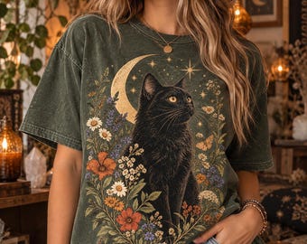 May include: A dark olive green t-shirt featuring a graphic print of a black cat sitting in front of a crescent moon. The cat is surrounded by a colorful arrangement of wildflowers, including daisies, orange poppies, and small purple blossoms.
