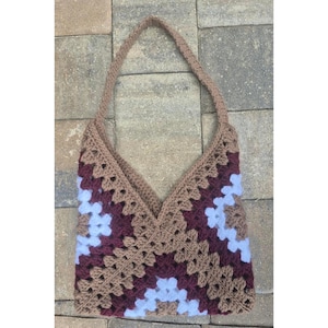 May include: A handmade crochet tote bag in a granny square pattern. The bag features a V-shaped opening and a shoulder strap. The color palette includes shades of brown, burgundy, and light blue, creating a unique and stylish accessory.