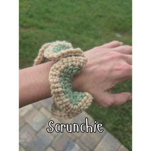 May include: A crocheted scrunchie in shades of tan and green, wrapped around a wrist. The scrunchie is handmade with a textured stitch, and the word "Scrunchie" is written in a decorative font at the bottom of the image.