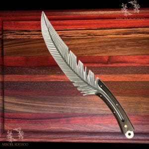 May include: A decorative knife with a feather-shaped blade. The silver blade features etched feather detailing. The handle is dark wood with silver accents. The knife is presented on a wooden chopping board.