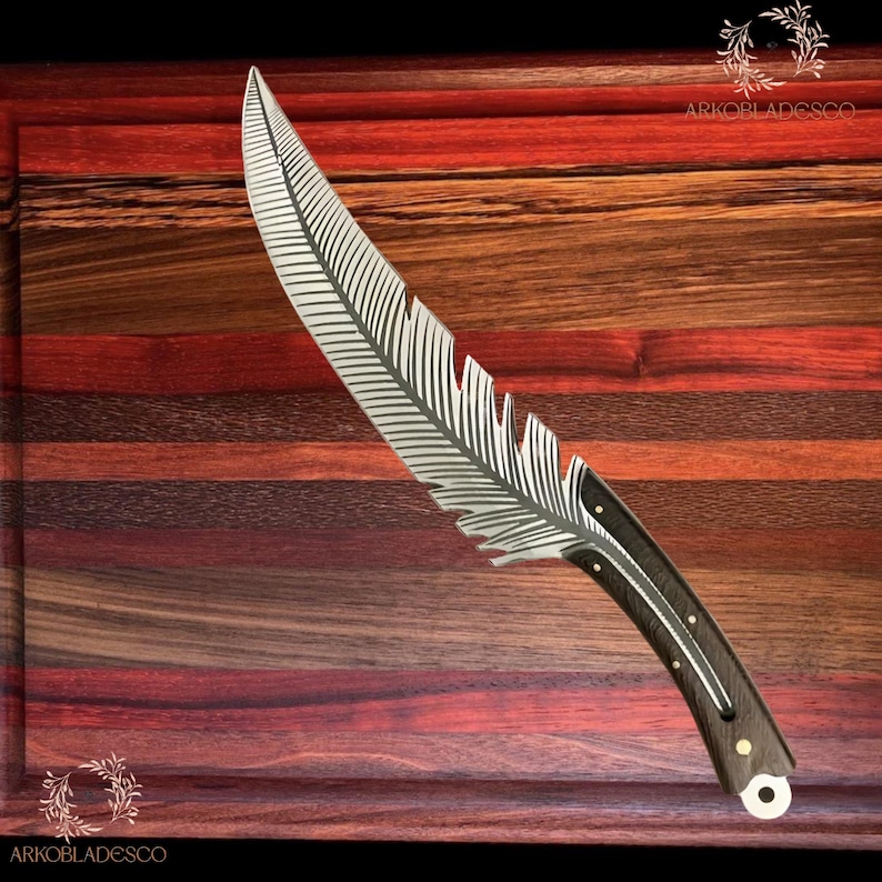 May include: A decorative knife with a feather-shaped blade. The silver blade features etched feather detailing. The handle is dark wood with silver accents. The knife is presented on a wooden chopping board.